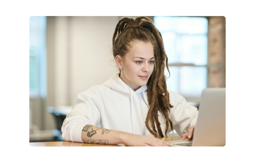 Young woman in a white hoodie working on a laptop, exploring creative income ideas in a modern co-working space.